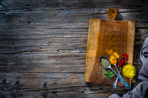 Culinary background: overhead view of herbs and spices arranged on a wooden cutting board at the right of a rustic wooden table background leaving useful copy space for text and/or logo at the left. High resolution 42Mp studio digital capture taken with SONY A7rII and Zeiss Batis 40mm F2.0 CF lens