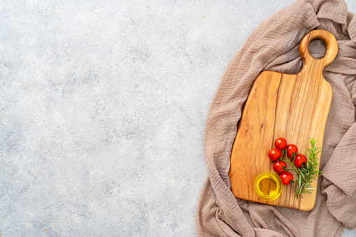 Culinary background: overhead view of a wooden cutting board with a brown textile tablecloth, cherry tomatoes, olive oil and rosemary. The composition is at the right of an horizontal abstract gray background leaving useful copy space for text and/or logo at the left. High resolution 42Mp studio digital capture taken with SONY A7rII and Zeiss Batis 40mm F2.0 CF lens