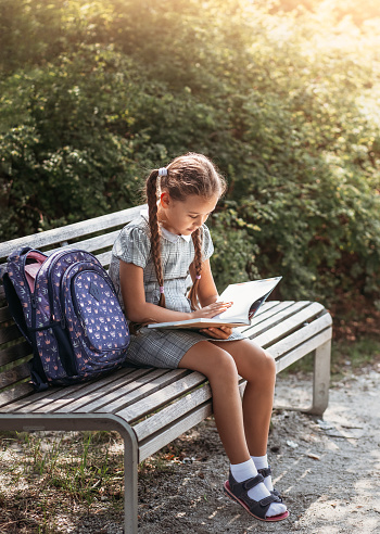 Girl with a backpack sitting on a bench with a book near the school. Back to school, lesson schedule, a diary with grades. Education, primary school classes, Back to school