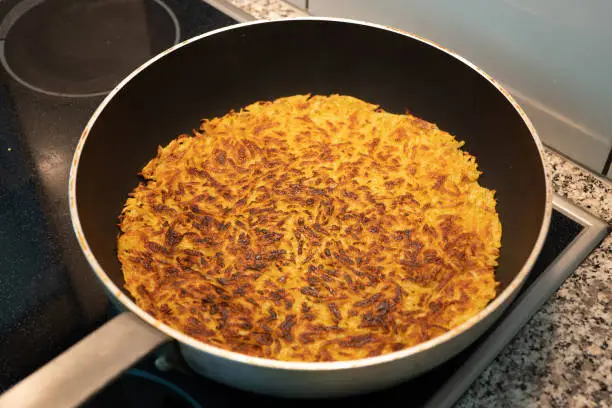 Homemade Swiss potato rosti or Rösti in a large frying pan on an electric stove. Top side view, no people.
