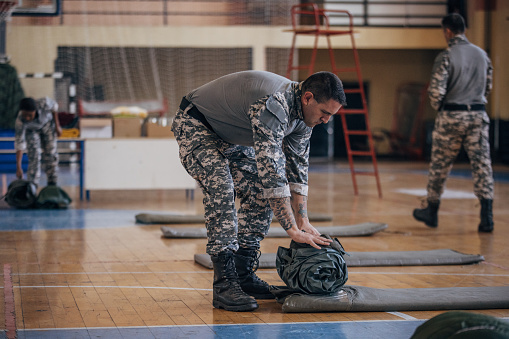 Soldiers on humanitarian aid preparing sleeping bags for civilians in school gymnasium, after natural disaster happened in city.