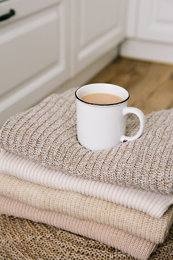 A cup of hot coffee with milk on a stack of knitted clothes. Autumn mood. The concept of home comfort, cozy mood and winter morning. Hygge. Vertical photo.