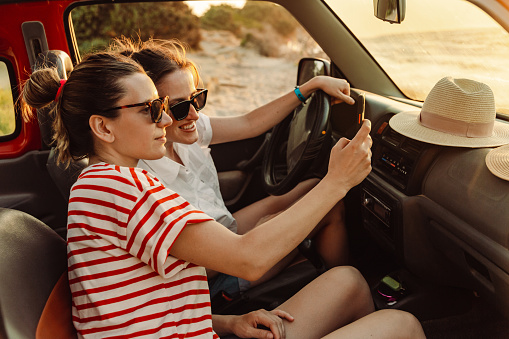 Two cheerful young women enjoying a summer’s road trip. One of them is driving a car and the other one holding a smart phone