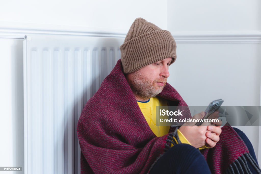 Cold man wearing blanket sitting next to radiator during cost of living crisis Portrait of a mid adult man wrapped up warm in layers of clothing, as well as a woolly blanket at woolly winter hat. He is sitting on the floor next to the radiator in his house, while looking at his cell phone. Room for copy space. Cold And Flu Stock Photo