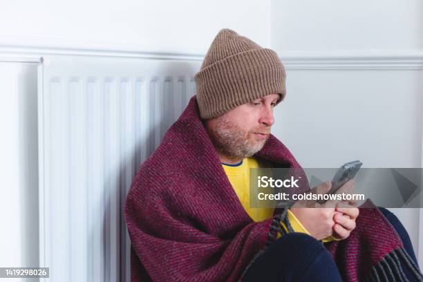 Cold Man Wearing Blanket Sitting Next To Radiator During Cost Of Living Crisis Stock Photo - Download Image Now