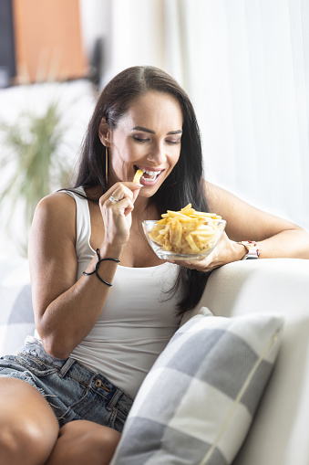 Smiling woman bites into one of french fries taken from a glass bowl while sitting at home on the sofa.