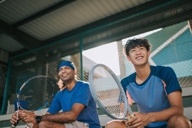 tenista profissional asiático sorrindo sentado em estádio observando companheiros de equipe praticando em quadra de tênis - warm light - fotografias e filmes do acervo