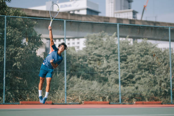 un joueur de tennis professionnel chinois d’origine asiatique sautant dans les airs en servant sur surface dure - tennis asian ethnicity male forehand photos et images de collection