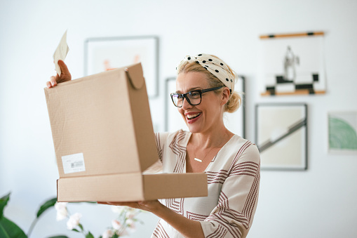 Close up shot of a beautiful blonde woman wearing glasses. She is standing, holding a carton box and a paper note from sender. She is smiling and looking excited to open it and see what is inside.