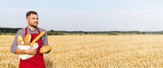 Baker stands in a wheat field and holds loaves of bread in his hands.