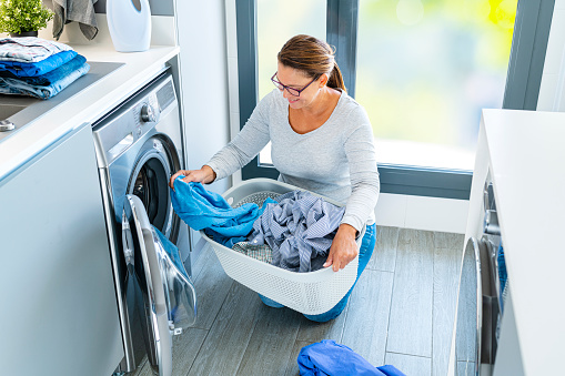 Mature smiling woman loading washing machine with laundry at home. Copy space available. High resolution 42Mp indoors digital capture taken with SONY A7rII and Zeiss Batis 40mm F2.0 CF lens