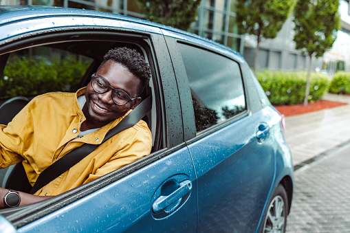 A smiling African-American man is holding the steering wheel and looking through the window on a rainy day