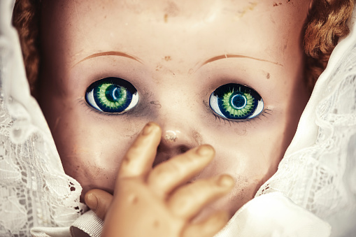 A close up of a spooky baby doll with green eyes covering her mouth with her hand.