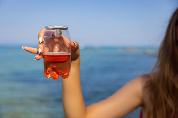 Unrecognizable young Caucasian woman  holding transparent plastic can with ice tea, with a sea as a background