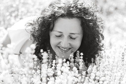 Black and white close-up portrait of curly-haired girl among flowers. Woman closed her eyes and enjoys aroma of lavender. Sincere smile. Digital detox, pleasure from slow life. Soft selective focus