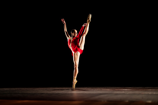 Girl dancing neoclassic ballet on dark stage wearing a red dress.