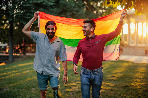 A bisexual male couple is outdoors with a rainbow flag in the park during sunset.