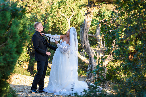 Black bride strangles her white husband's tie against the backdrop of a sunny forest