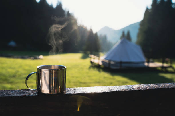 closeup of a steaming metallic cup with warm coffee or tea in the morning with glamping camping ground in the background stock photo