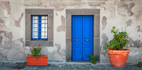Front view and close up of old gray plaster wall with double blue door and window. Potted plants in front of the house as decoration. Naples, Italy.