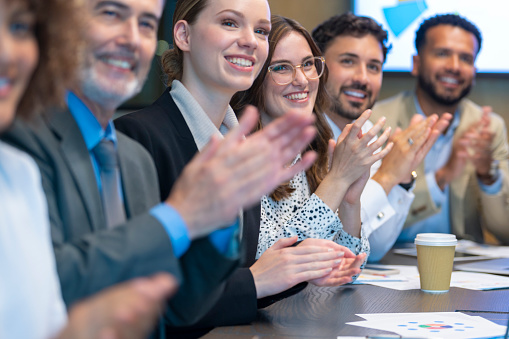 Group of business people applauding a presentation. They are sitting at a conference table. There are several ethnicities present including Caucasian, African and Latino