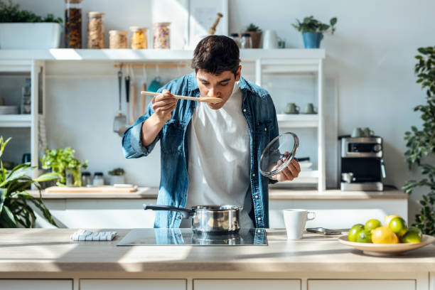 bel homme mûr goûtant la nourriture qu’il prépare dans la cuisine à la maison. - cooking chef domestic kitchen food photos et images de collection