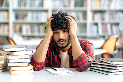Upset student of Arabian or Indian nationality in stylish clothes, sits at a desk in the library, tired of preparing for the exam, reading books, feels exhausted and tired, needs rest