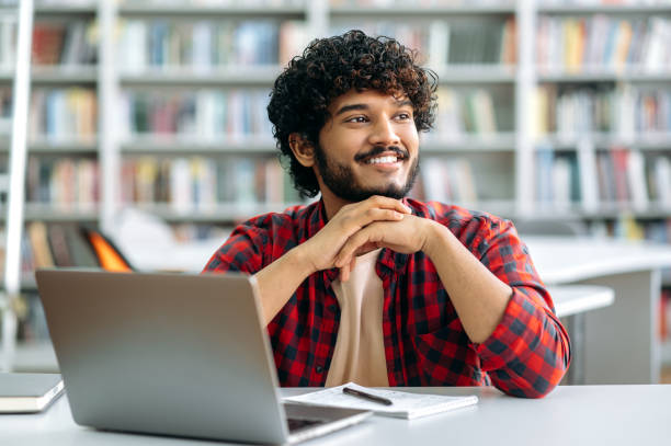 istock Positive attractive curly-haired Indian or Arabian smart guy, student or freelancer, dressed in trendy casual clothes, sits at a desk with laptop in the library, looks away, smiling friendly 1419416567