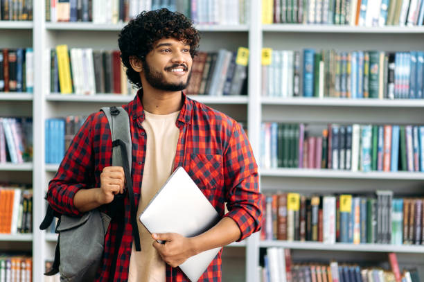 pozytywny pewny siebie indyjski lub arabski student uniwersytetu, w stylowym stroju na co dzień, z plecakiem i laptopem, stoi w bibliotece na tle półek z książkami, odwraca wzrok, uśmiecha się, marzy - college student student asian ethnicity high school student zdjęcia i obrazy z banku zdjęć