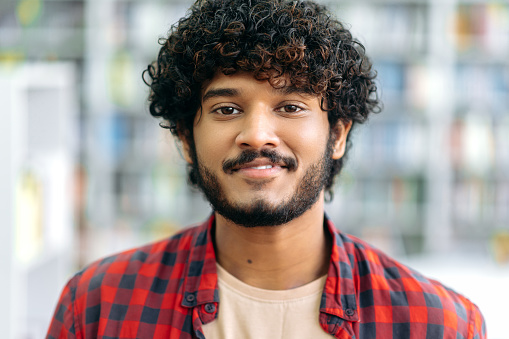 Close-up photo of a handsome positive curly haired indian or arabian guy, in casual shirt, manager, designer, student or freelancer, stands indoors, looks at camera, smiling friendly