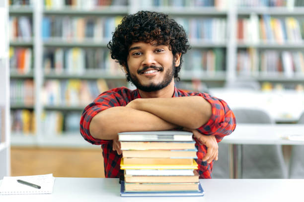 istock Positive clever indian or arabian guy, mixed race male, university student, sits at a desk with books, in the library against the background of bookshelves, looks at camera, smiles friendly 1419416607