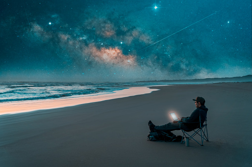 mature latin male sitting at night on the shore of the beach with a laptop and stars in the background