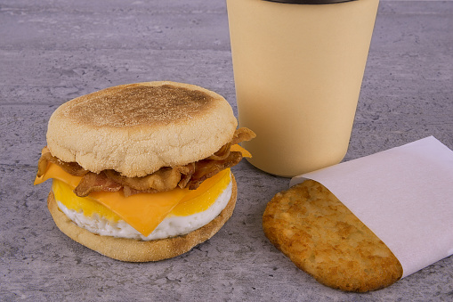 Breakfast sandwich with coffee and hash brown on concrete table. English muffin, egg, cheese and sausage.