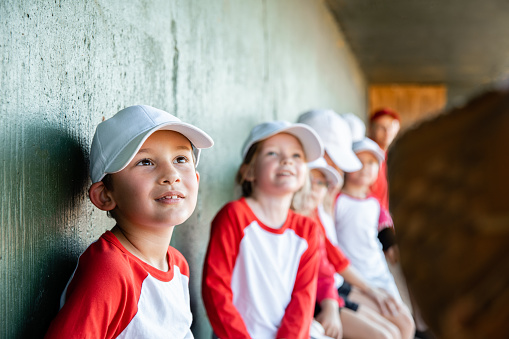 Elementary age children are athletes playing little league baseball together on co-ed sports team with coaches at ballfield. Teammates are wearing matching uniforms and baseball caps.