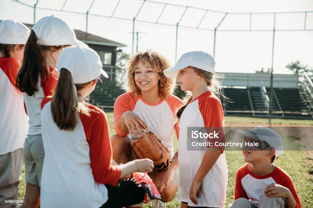 Elementary age children are athletes playing little league baseball together on co-ed sports team with coaches at ballfield Elementary age children are athletes playing little league baseball together on co-ed sports team with coaches at ballfield. Teammates are wearing matching uniforms and baseball caps. Coach Stock Photo