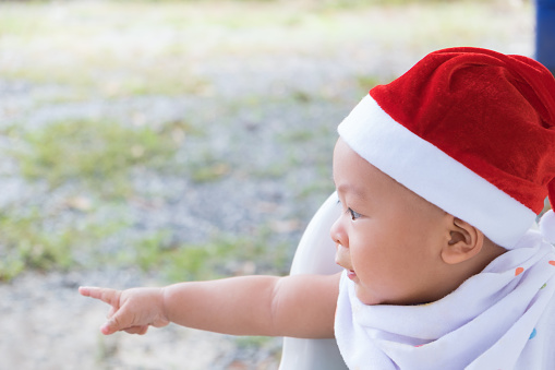 Happy little Asian baby boy in red Santa Claus hat celebrates Christmas while sitting on chair at outdoors. The kid pointed his finger out in front of the New Year holiday. Breastfeeding early childhood development.