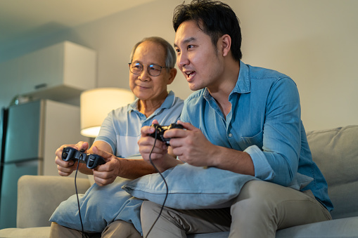 Asian senior father and handsome son play video game together at home. Attractive senior older and young boy sit and holding game controller feeling excited and fun with smiling face in living room.