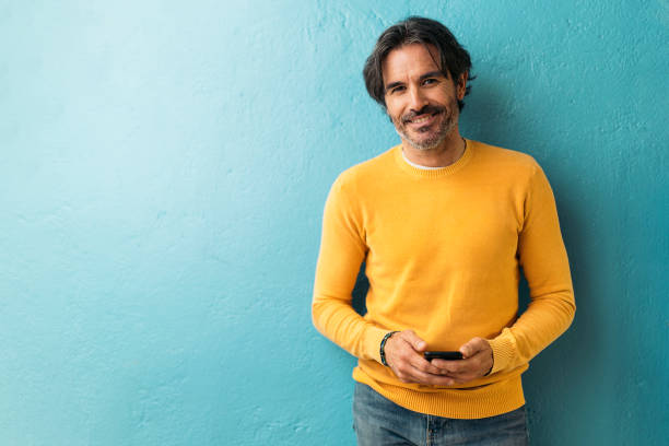 Mature Business Man Portrait Smiley male office worker holding his mobile phone and looking at camera against blue background. 45 49 years stock pictures, royalty-free photos & images