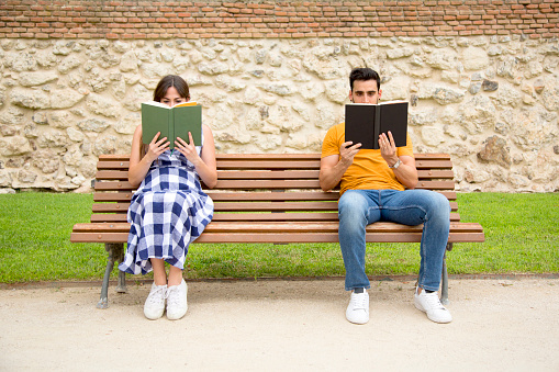 Frontal view of two people sitting on a bench of a park reading books