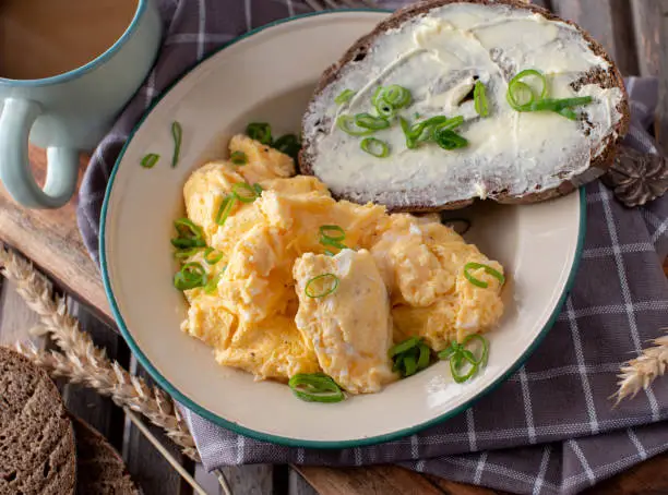 Rustic and rural breakfast plate, old fashione style with homemade scrambled eggs and buttered sourdough bread. Served with a cup of coffee on wooden background