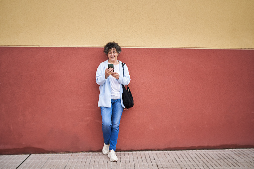 Full length view of a mature woman in jacket and jeans holding her purse leaning against a two-color wall outdoors using her cell phone.