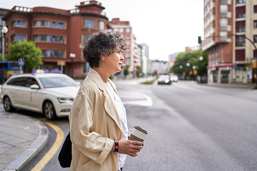 Side view of a serious mature woman crossing the street while looking at the traffic light holding a reusable coffee mug.