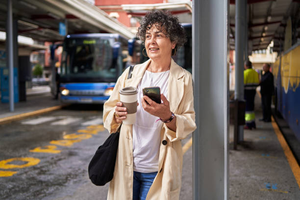 eine reife frau, die an einem busbahnhof wartet und ihr handy und ihre tasse kaffee hält, während sie wegschaut - pay phone telephone people women stock-fotos und bilder