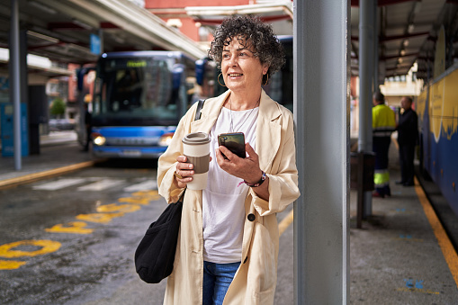 Portrait of a smiling mature woman leaning on a metal column waiting at a bus terminal holding her phone and cup of coffee while looking away.