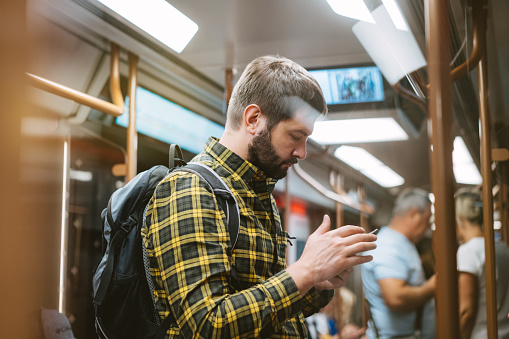 caucasian man in checkered plaid shirt with backpack traveling by metro, standing in subway car holding mobile telephone in hands.Image with selective focus.