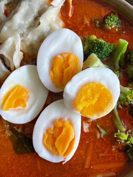 Full frame image of bowl of home made, spicy coconut milk laksa soup with wontons (Chinese dumplings), broccoli, thick wheat noodles and boiled egg halves, elevated view stock photo