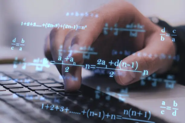 Close up of businessman hands using laptop keyboard at workplace with monitor, supplies and glowing mathematical formulas on blue background. Education, knowledge and statistics concept