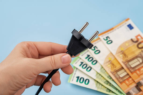 Woman hand holds electric plug against fan shaped euro banknotes over blue background. Energy price spike in Europe. Electric bill increases. Soaring of electricity prices concept. Top view. la mano de la mujer sostiene el enchufe eléctrico contra los billetes de euro en forma de ventilador sobre fondo azul. repunte de los precios de la energía en europa. aumenta la factura eléctrica. alza del concepto de precios de la electricidad.