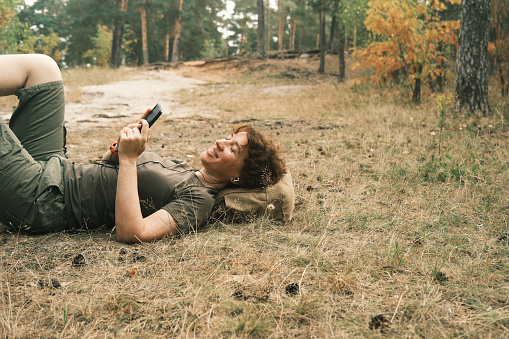 Cheerful mature woman is lying on ground in autumn forest and listening to the music by headphones