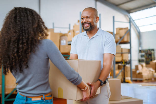 Shipping, logistics and delivery worker help a woman customer with a box in a courier warehouse. Stock and cargo man working in a supply chain factory with a happy smile and hands over package stock photo